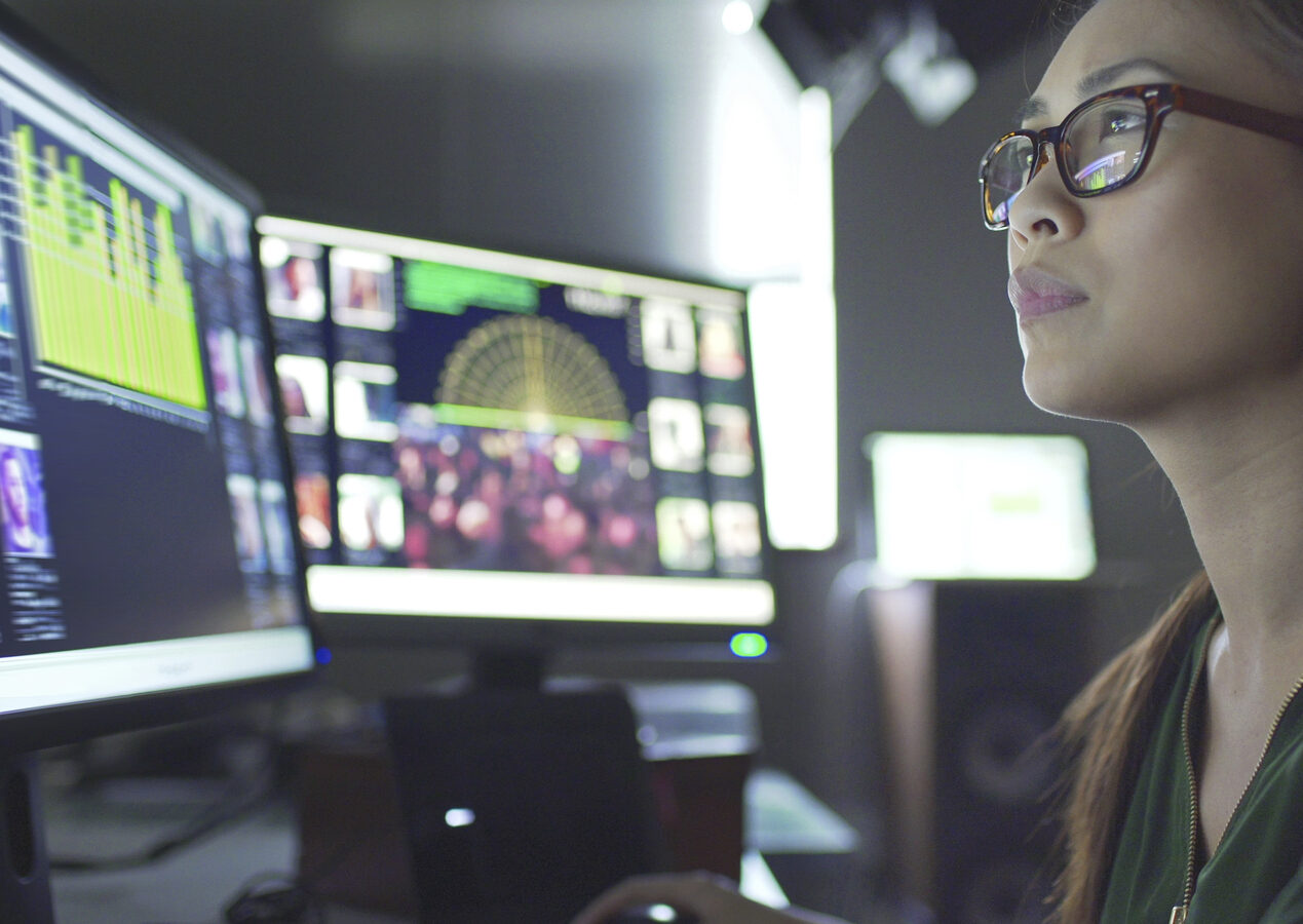 Close up stock image of a young asian woman sitting down at her desk where she’s surrounded by 3 large computer monitors displaying out of focus images of people as thumbnails; crowds; graphs & scrolling text.